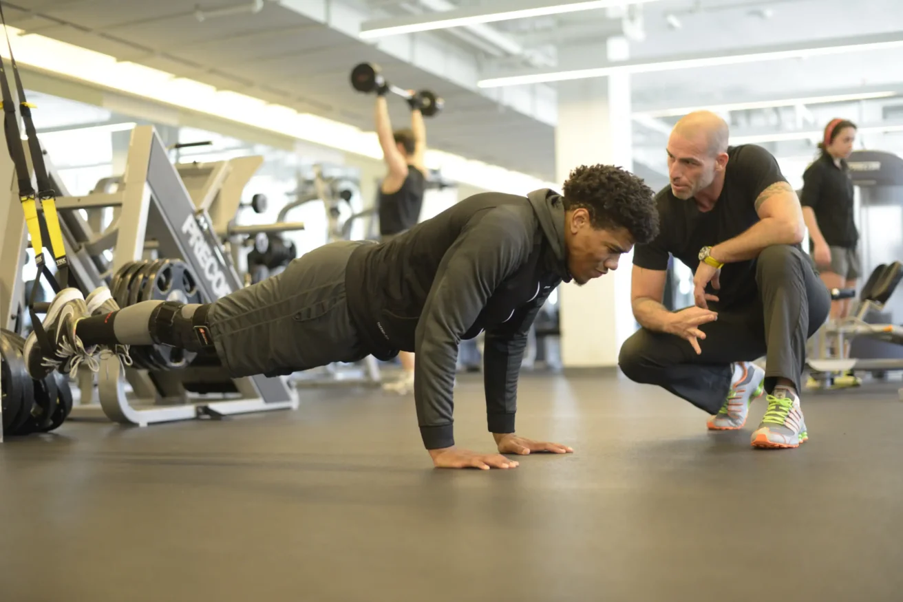 A trainer crouches and gives instructions to a man doing push-ups in a gym, with other people exercising in the background.