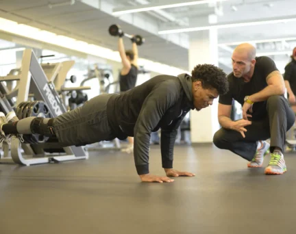 A trainer crouches and gives instructions to a man doing push-ups in a gym, with other people exercising in the background.
