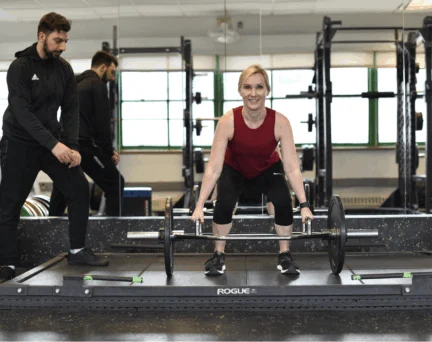 A woman in athletic attire performs a barbell deadlift in a gym while an Asphalt Green trainer provides guidance on form.