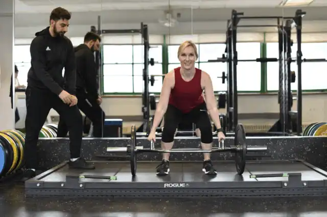 A woman in athletic attire performs a barbell deadlift in a gym while an Asphalt Green trainer provides guidance on form.