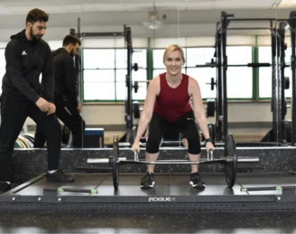 A woman in athletic attire performs a barbell deadlift in a gym while an Asphalt Green trainer provides guidance on form.