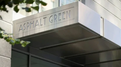Sign reading "Asphalt Green" on a metal awning attached to a modern glass and stone building, with leaves slightly visible in the foreground.