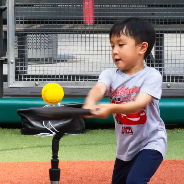 A young boy hits a yellow ball off a baseball tee on a field, with bleachers and a metal fence in the background.