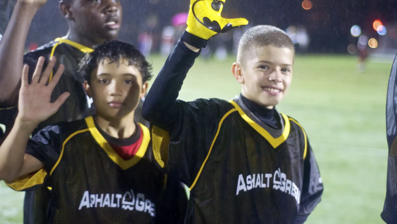 Three boys in “Asphalt Green” jerseys stand outdoors, two raising their hands, one wearing a yellow sports glove, with blurred lights in the background on a rainy night.