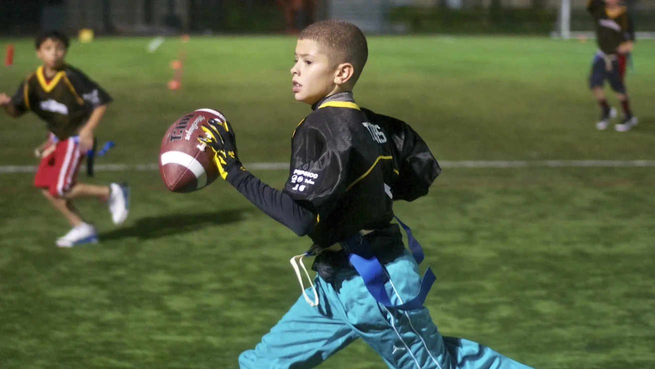 A boy in a black jersey and blue pants runs on a field holding a football, with other children playing flag football in the background.