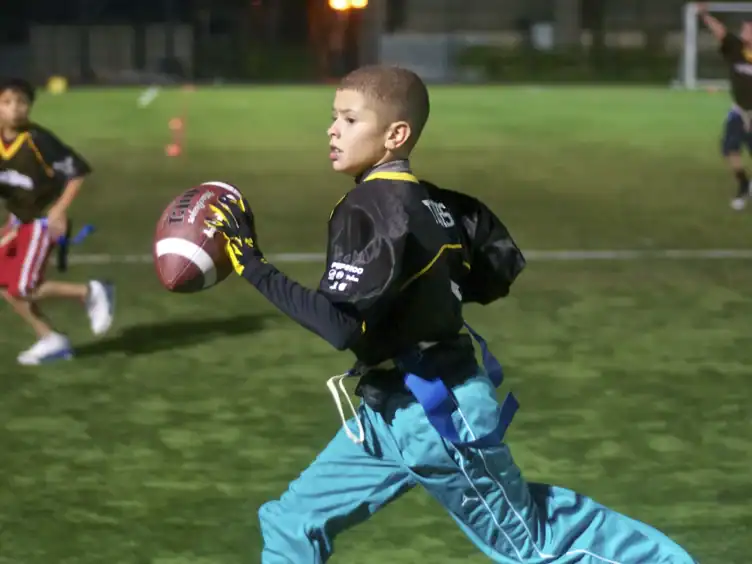 A boy in a black jersey and blue pants runs on a field holding a football, with other children playing flag football in the background.