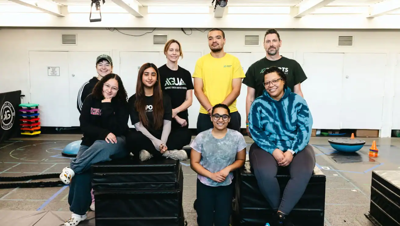 Eight people pose indoors in a gym or training room, some seated on black mats and others standing behind them, with exercise equipment visible in the background.