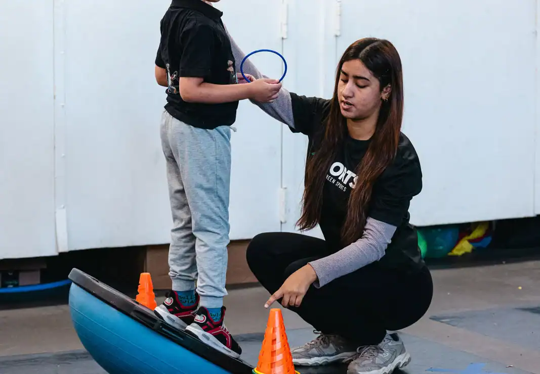 A child stands on a balance trainer holding a ring while an adult kneels beside him, pointing at orange cones on the floor.