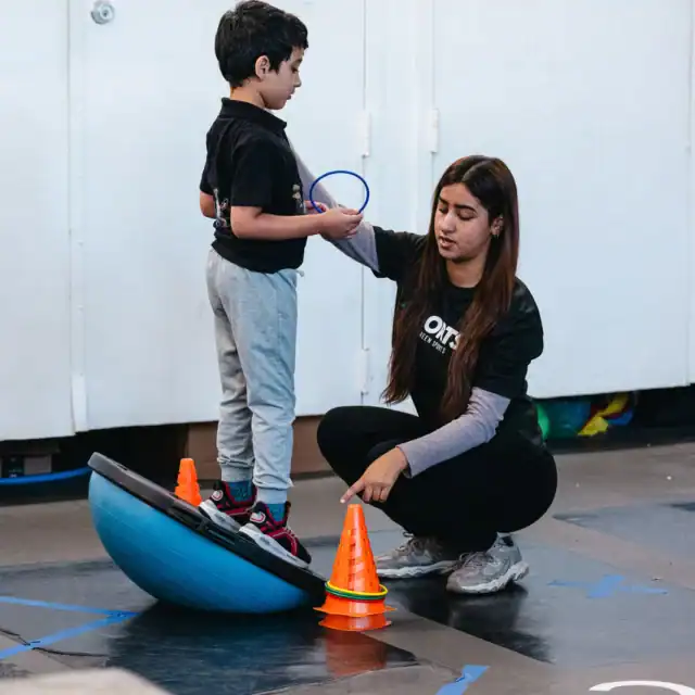 A young boy stands on a balance trainer holding a paddle, while a woman kneels beside him, pointing to orange cones on the floor in a gym setting.