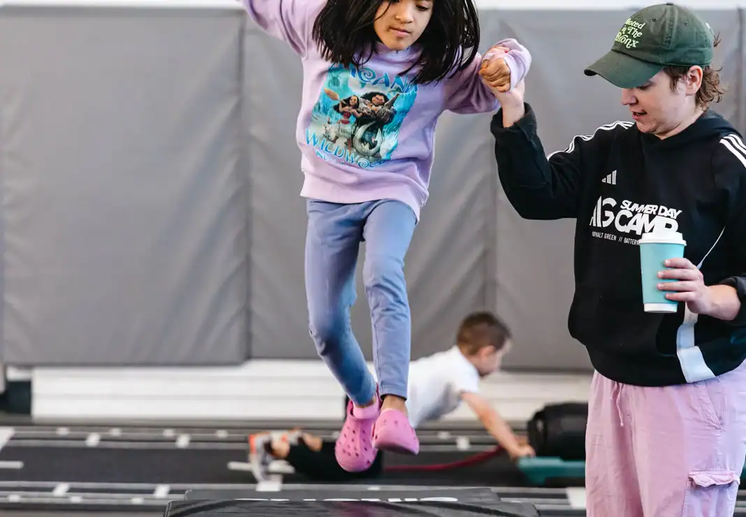 A young girl jumps onto a padded platform with assistance from an adult in a gym setting. Another child is visible in the background.