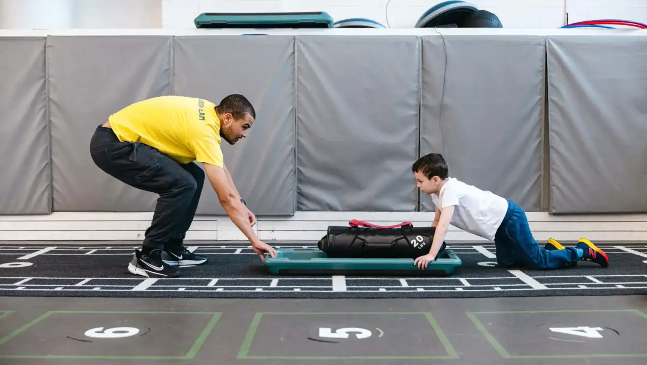 An adult and a child face each other in a gym, both crouched down; the child pushes a weighted bag on a board across the floor while the adult supervises.