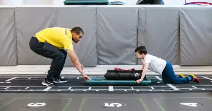 An adult and a child face each other in a gym, both crouched down; the child pushes a weighted bag on a board across the floor while the adult supervises.