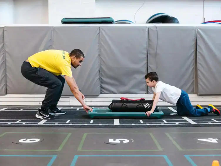 An adult and a child face each other in a gym, both crouched down; the child pushes a weighted bag on a board across the floor while the adult supervises.