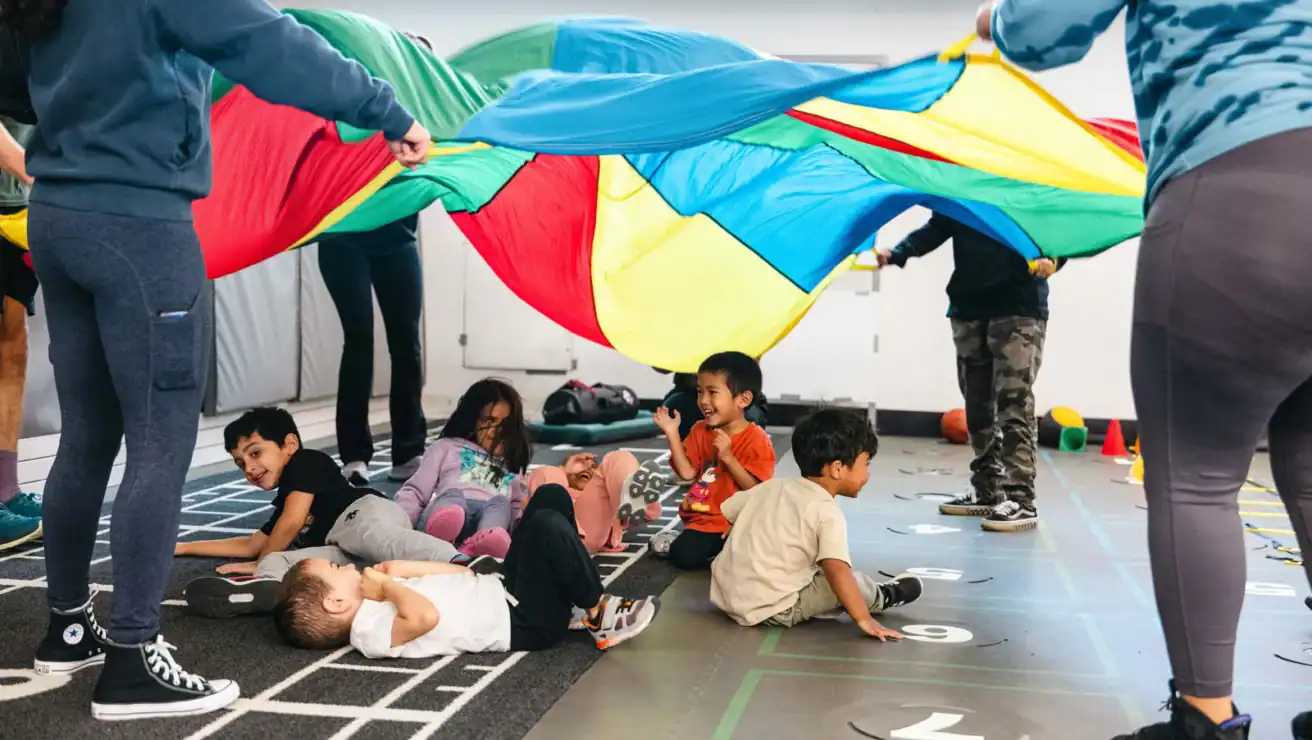 Children sit and lie on the floor while adults hold up a colorful parachute above them in a gym or play area.