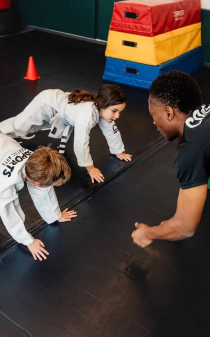 A martial arts instructor instructing two children who are holding planks on a black mat.