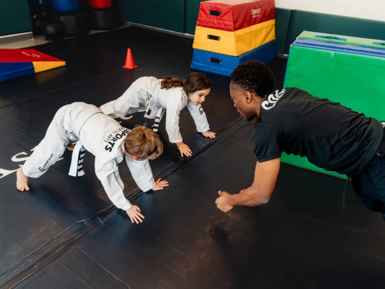 A martial arts instructor instructing two children who are holding planks on a black mat.