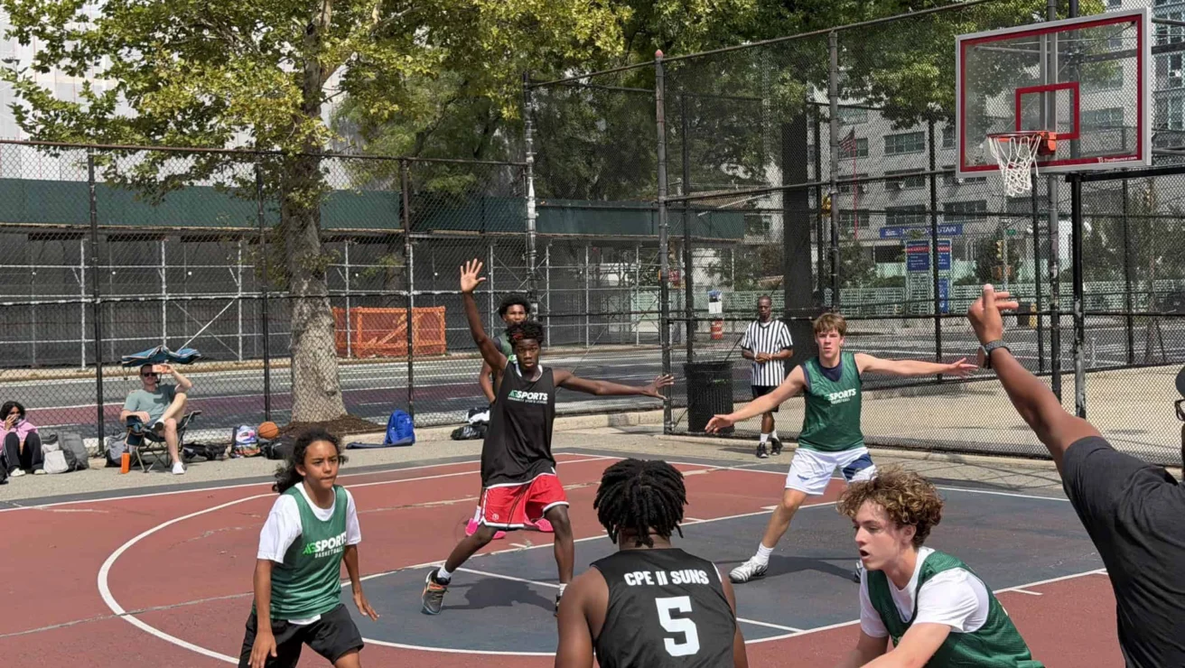 A group of teens play a competitive basketball game on an outdoor court, with two teams in black and green jerseys actively defending and attacking near the basket.