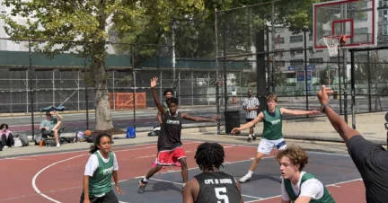A group of teens play a competitive basketball game on an outdoor court, with two teams in black and green jerseys actively defending and attacking near the basket.