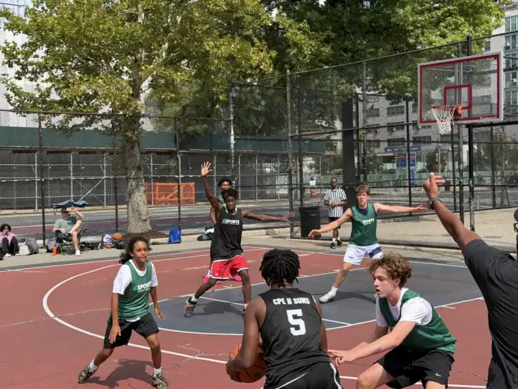 A group of teens play a competitive basketball game on an outdoor court, with two teams in black and green jerseys actively defending and attacking near the basket.