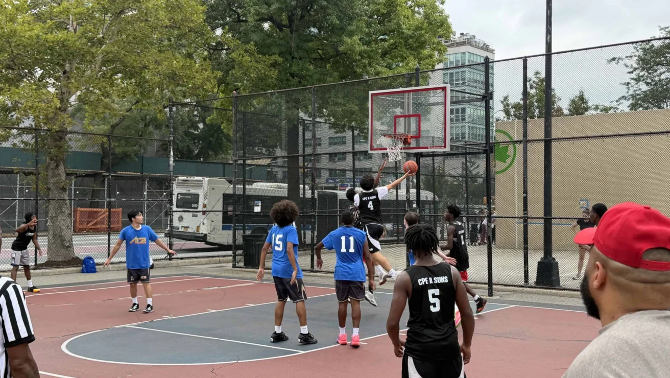 A player in a black jersey leaps for a layup during an outdoor basketball game, while players in blue and black jerseys watch near the hoop. Spectators and referees stand nearby.