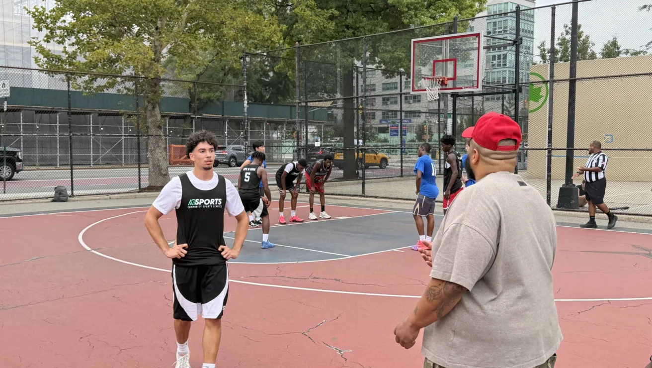 A group of people play basketball on an outdoor court. One player stands near the sideline while others prepare for action under the hoop. A coach or spectator stands nearby.