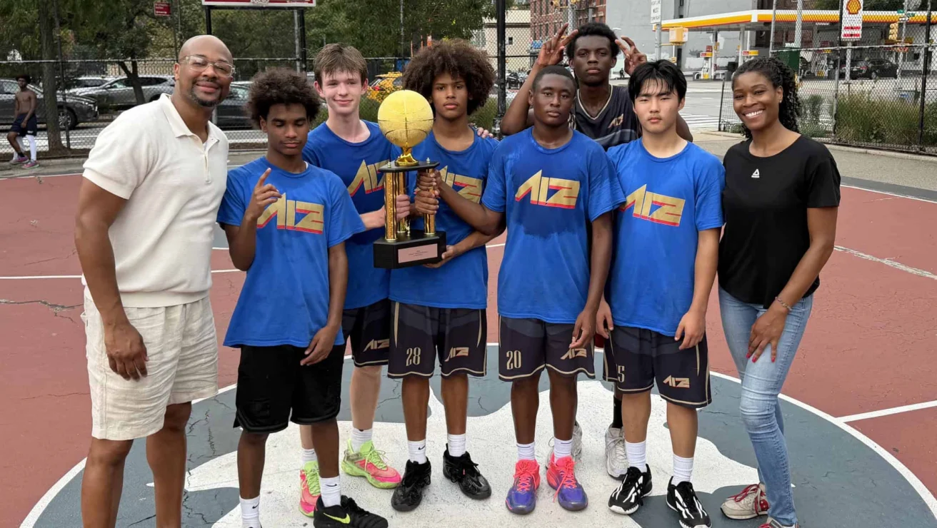 A boys’ basketball team in blue shirts poses with a trophy and two adults on an outdoor court, with trees and city buildings in the background.