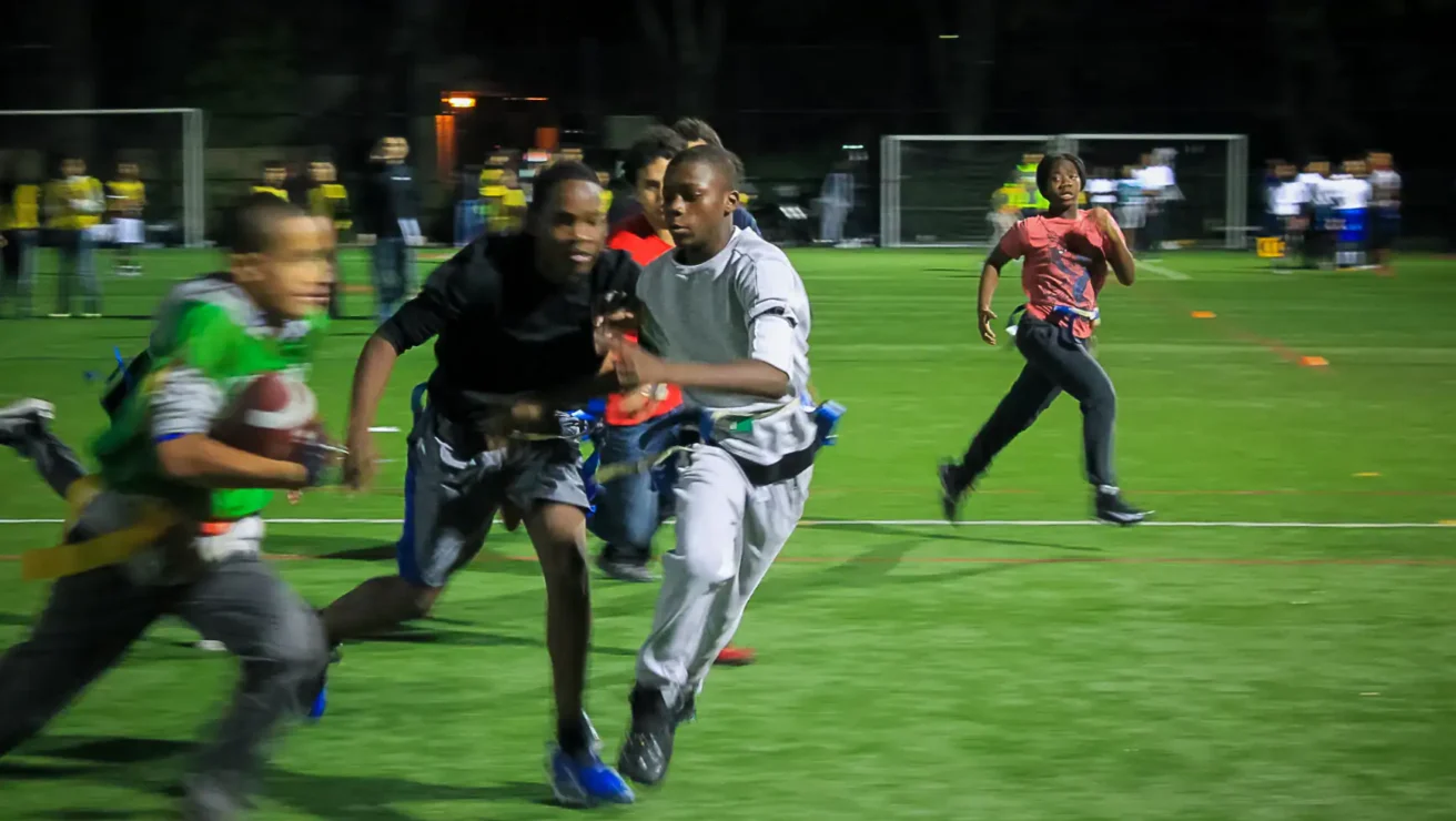 Four boys play flag football at night on a turf field, with one running with the ball as others chase him; blurred motion indicates fast movement.