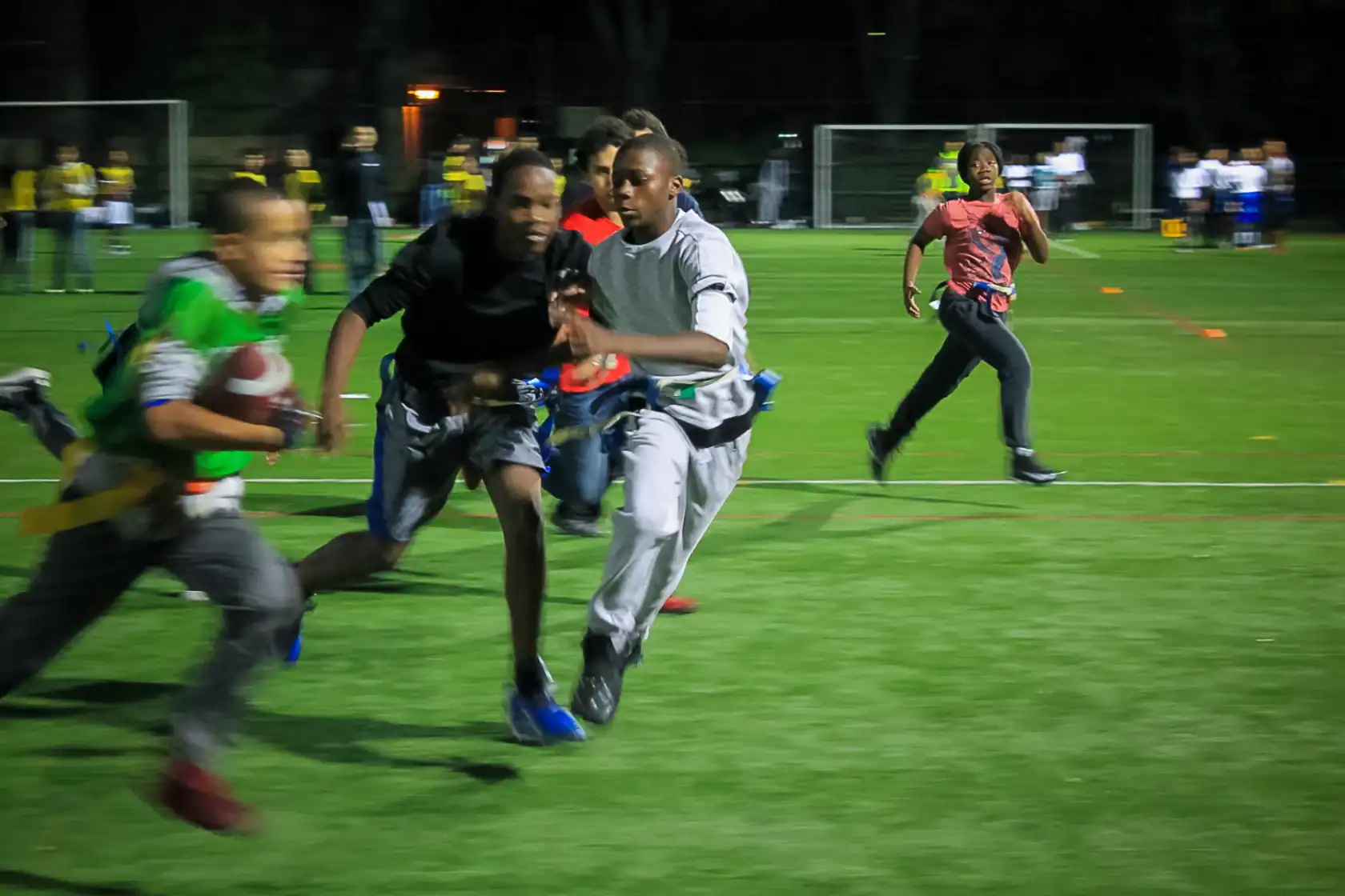 Four boys play flag football at night on a turf field, with one running with the ball as others chase him; blurred motion indicates fast movement.