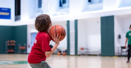A child in a red shirt prepares to shoot a basketball in an indoor gym.