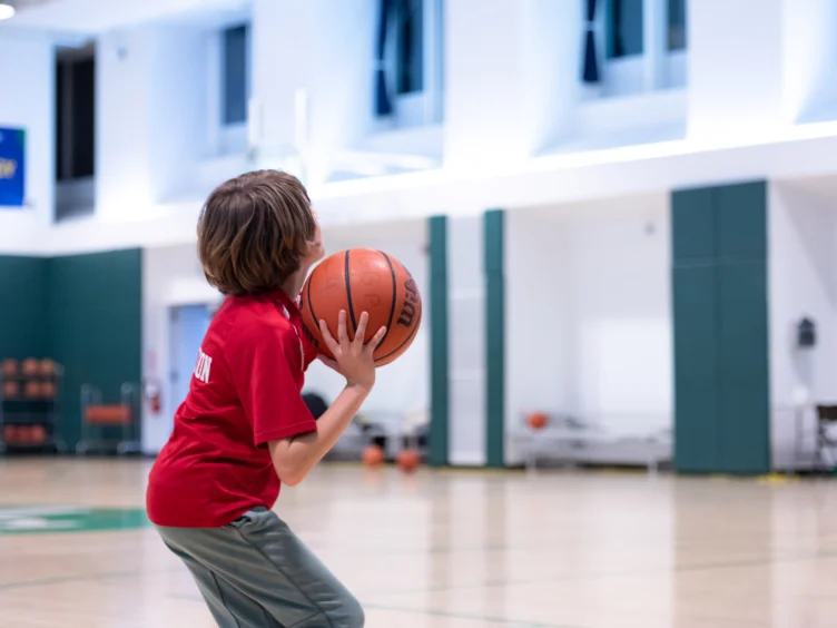 A child in a red shirt prepares to shoot a basketball in an indoor gym.