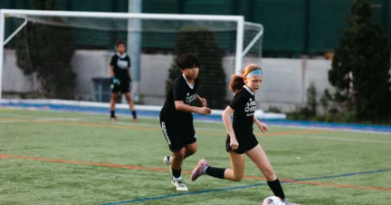 Two young soccer players in black uniforms run on a field, with a goalpost in the background.