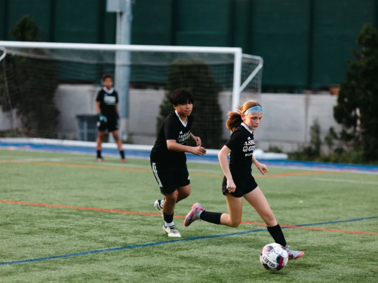 Two young soccer players in black uniforms run on a field, with a goalpost in the background.