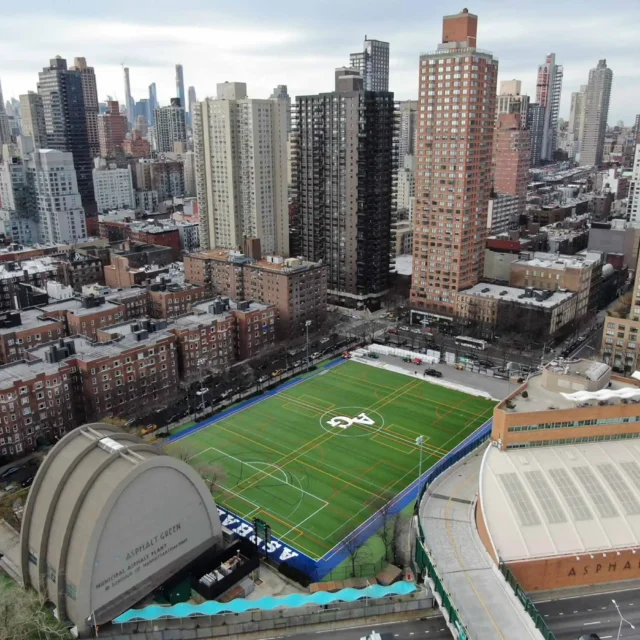 Aerial view of a cityscape featuring a fenced green sports field and surrounding buildings.