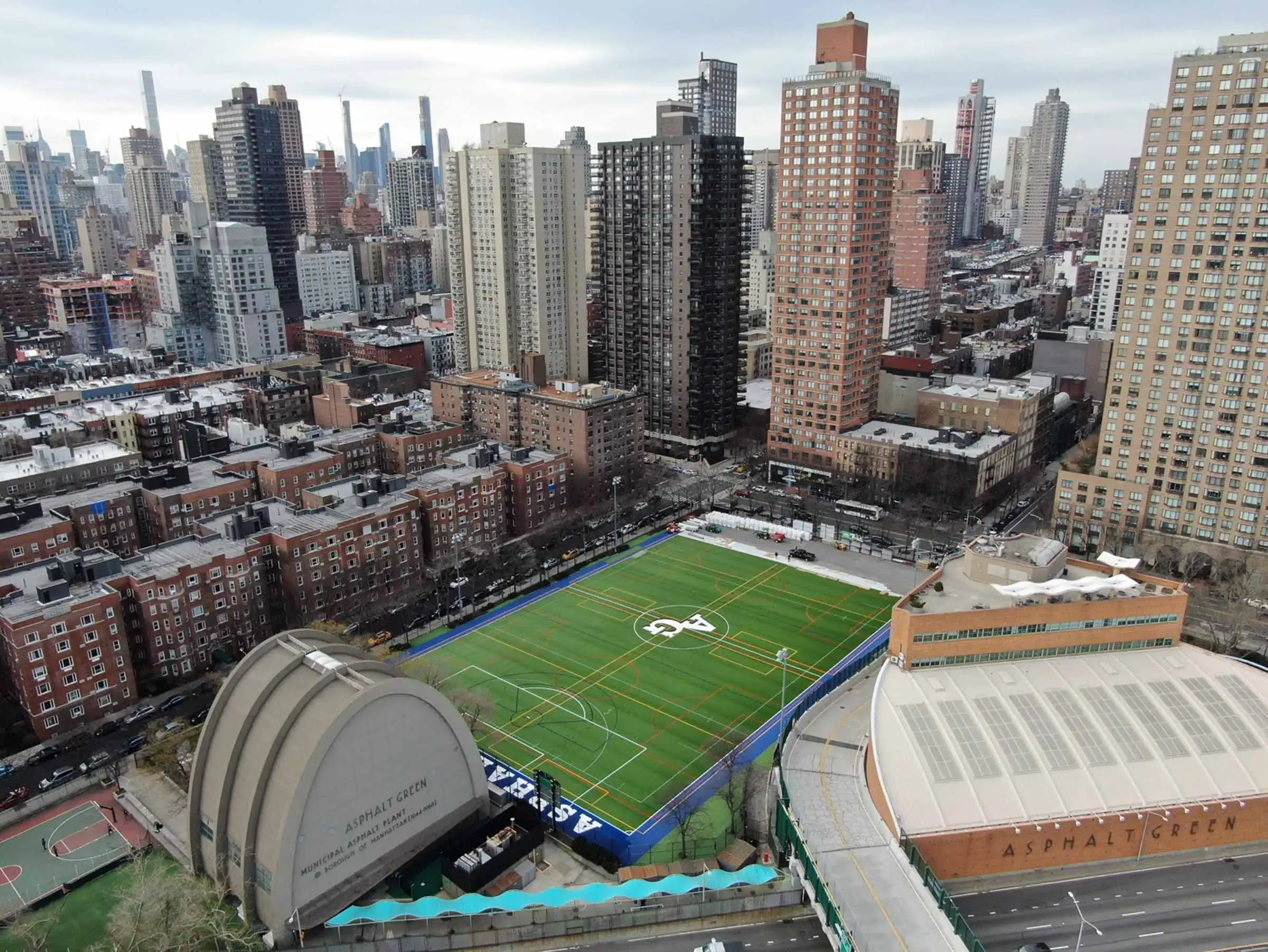 Aerial view of a cityscape featuring a fenced green sports field and surrounding buildings.