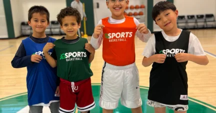 Four children in sports clothing, smiling and standing on a basketball court. They each wear a jersey with the word "Sports" on it.
