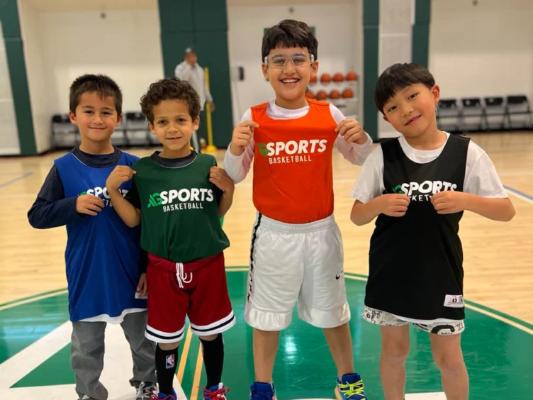 Four children in sports clothing, smiling and standing on a basketball court. They each wear a jersey with the word "Sports" on it.