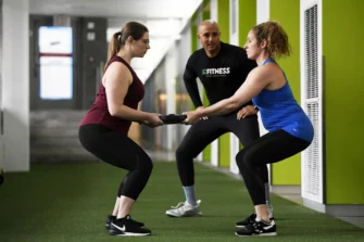 Two women squat while holding a weight plate together, guided by a fitness instructor in a gym setting.
