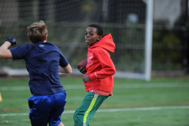 Two boys playing football on a field, one in a red hoodie holding the ball, and the other in a blue shirt running nearby.