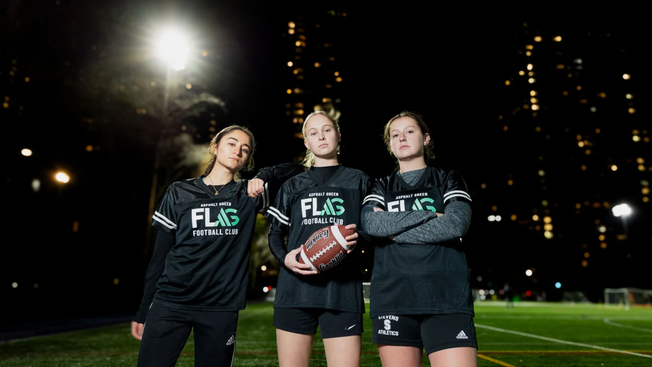Three people in matching uniforms stand on a lit sports field at night, holding a football, with city lights in the background.