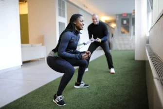 A woman performs squats with a resistance band, assisted by a man in a hallway with artificial turf flooring.