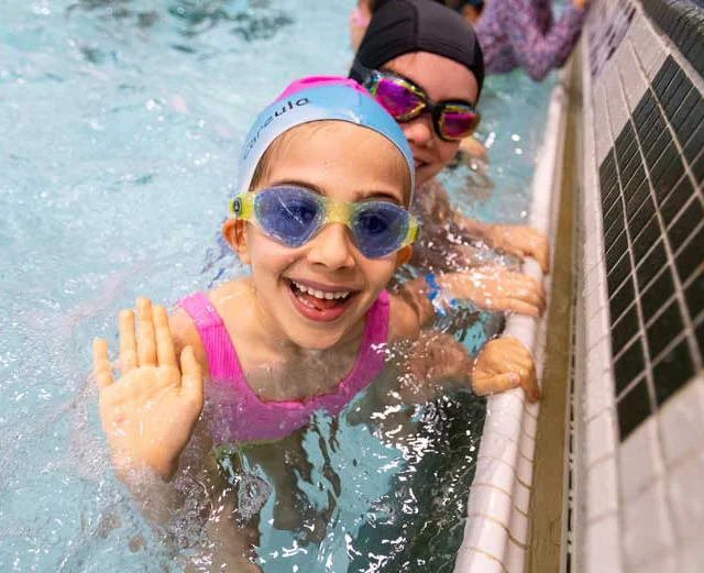 A smiling child wearing goggles and a swim cap waves from a pool, with another person partially visible in the background.