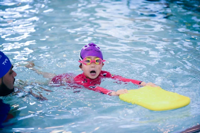 Child in a purple cap and goggles practices swimming with a yellow kickboard in a pool. An adult wearing a swim cap is nearby.