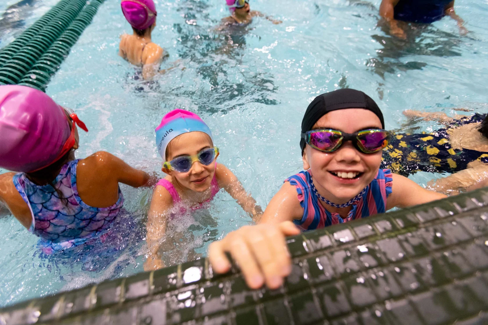 Children in swim caps and goggles smiling at the edge of an indoor pool.