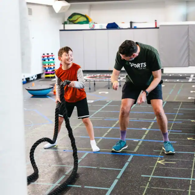 A young boy and an adult exercise together indoors using battle ropes on a gym floor marked with colorful lines and shapes.