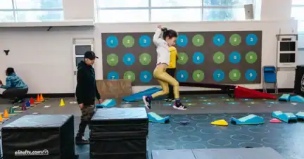 A child jumps off a black foam block in an indoor gym with various soft play equipment, while another child watches nearby.
