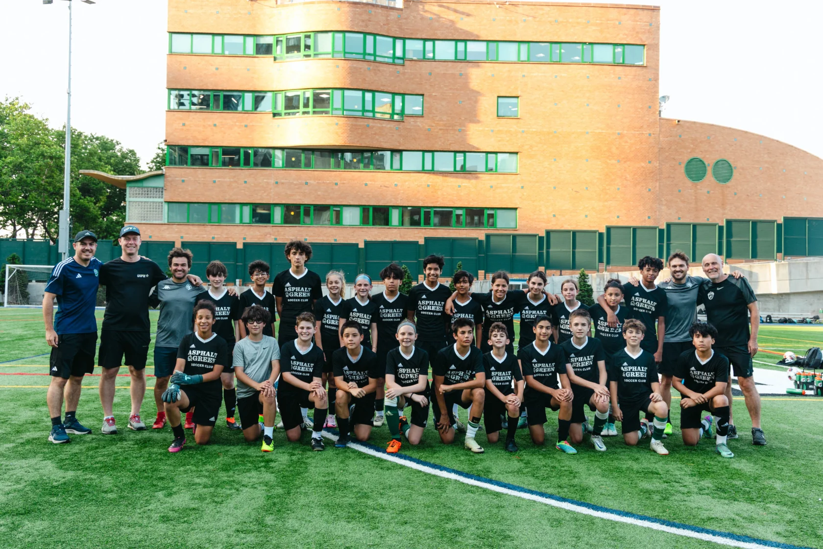 A soccer team poses for a group photo on a field, with coaches standing on either side. A brick building is in the background.