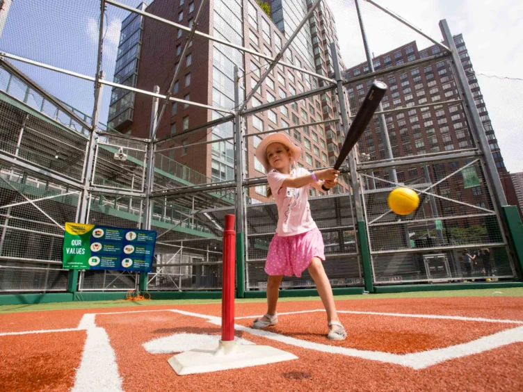A young girl in a pink outfit and hat swings a bat at a stationary ball on a tee in an outdoor urban baseball field.