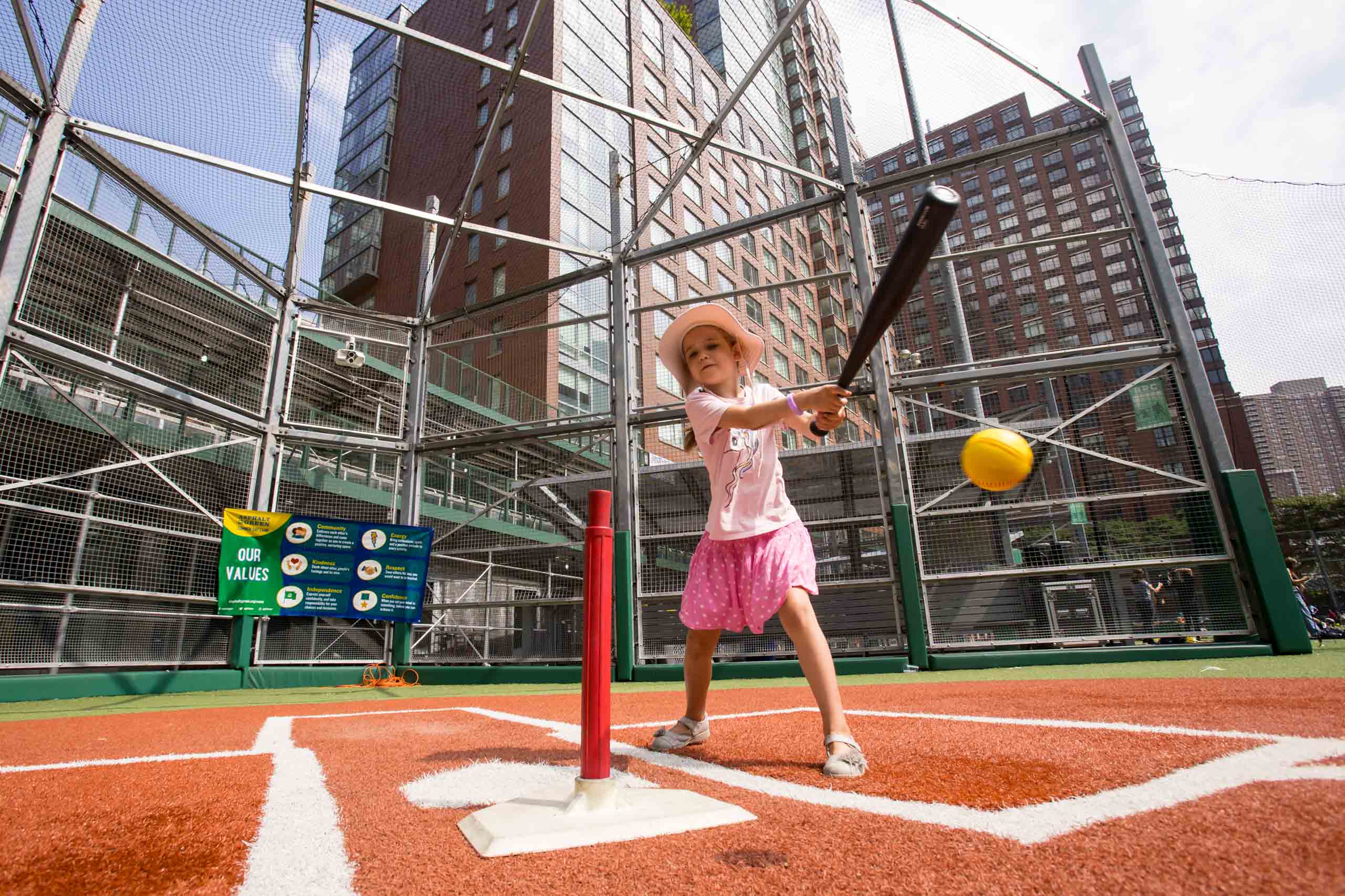A young girl in a pink outfit and hat swings a bat at a stationary ball on a tee in an outdoor urban baseball field.