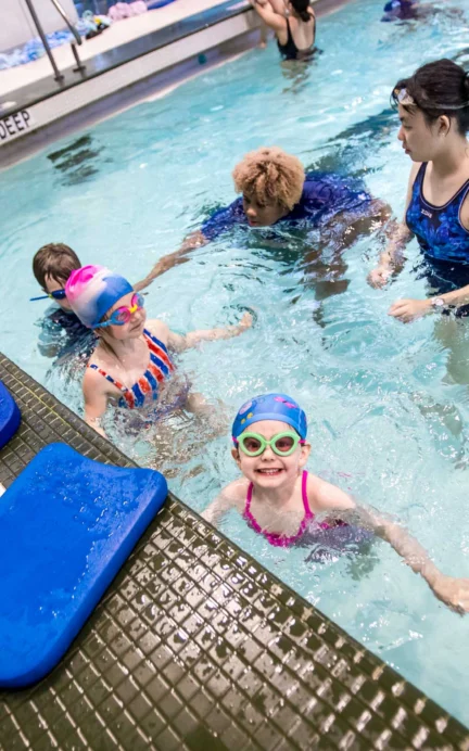 Children in a swimming pool during a lesson, guided by two instructors. Brightly colored swim gear is visible, and pool depth markings are shown.