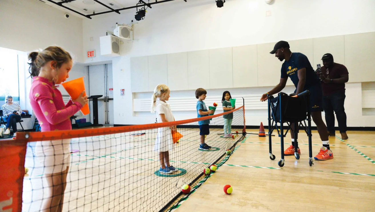 Children participate in an indoor tennis lesson, standing on one side of a net. An instructor assists them using a walker. Tennis balls are scattered on the floor.
